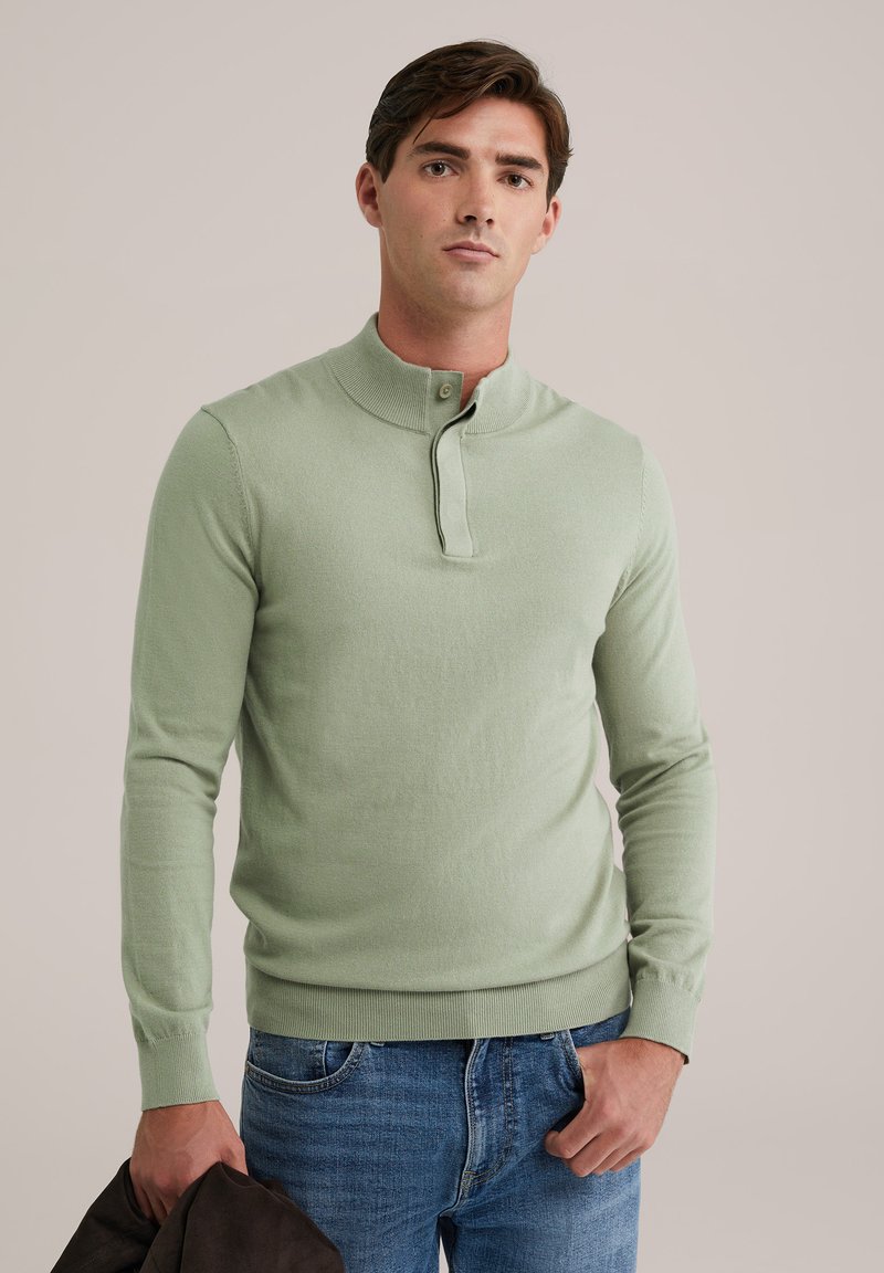 Gai zaļš, garām piedurknēm adīts džemperis ar ribinātu apkakli un pogu aizdari. Materiāls izskatās mīksts un ar pieguļošu formu. Vilkts ar ziliem džinsiem.