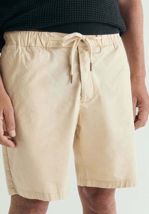 Pantalones cortos beige hasta la rodilla con cintura elástica y cordón anudado, usados por una persona con una camiseta negra, manos relajadas a los costados.