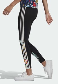 Černé legíny se třemi bílými pruhy po straně, na spodním lemu s květinovými vzory v jasných barvách. Nohavice jsou doplněny bílými teniskami.