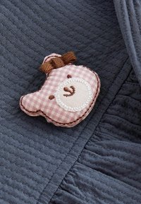 Écusson en tissu vichy rose représentant un visage d'ours avec détails brodés et nœud tricoté marron sur un tissu matelassé bleu foncé texturé.
