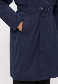 Marinblå trench coat med knytband i midjan, dubbelknappstängning och sidofickor. Tillverkad av ett slätt, vattenavvisande material med en figursydd passform.