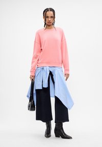 Korallfarget genser med vide ermer, lagd over en blåstripet skjorte, kombinert med mørke vide bukser og svarte ankelstøvler.