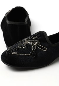 Zapatos tipo slip-on de terciopelo negro con un diseño detallado de spider de cuentas en negro y acentos metálicos. Textura suave y puntera redondeada.