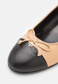 Bailarinas con una parte superior beige y puntera negra. Cuentan con una decoración de lazo y un detalle en dorado. Textura suave en toda su superficie.