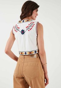White sleeveless crop top with colorful floral embroidery on the back and hem. Worn with tan high-waisted pants, showcasing a fitted design.