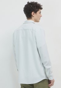 Jeune homme de profil à droite portant une chemise à manches longues blanche avec de fines rayures verticales et un pantalon vert olive sur fond uni.