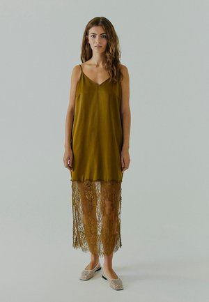 Oliven grøn glidedragt med justerbare stropper, der har en lang blonde kant. Glat tekstur med detaljeret blonde ved bunden.