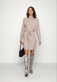 Strickkleid in Beige mit langen Ärmeln und Bindegürtel, kombiniert mit knielangen, gewellten Stiefeln in Schwarz-Weiß-Muster und einer schwarzen Tasche.