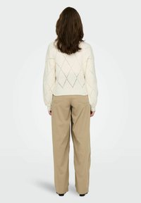 Femme aux cheveux bruns mi-longs portant un pull blanc à motifs en losange et un pantalon beige à jambes larges, se tenant de dos.