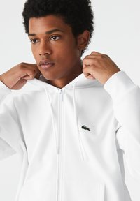 Jeune homme ajustant la capuche d’un sweat zippé blanc avec un petit logo crocodile vert sur la poitrine.