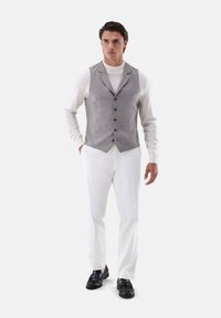 Grau strukturierte Weste mit fünf Knöpfen, getragen über einem weißen Rollkragenpullover, kombiniert mit weißen Hosen und schwarzen Schuhen. Schlichtes Design und taillierte Passform.