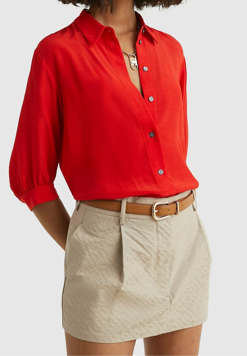 Femme portant un chemisier rouge à manches bouffantes, boutonné et rentré dans une jupe beige à motifs, ceinturée d'une ceinture en cuir marron clair à boucle dorée.