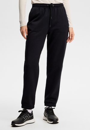 Persona che indossa pantaloni jogger neri con polsini elastici e scarpe da ginnastica nere e bianche, in piedi su uno sfondo neutro.