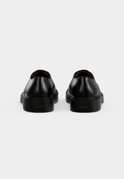 Paire de chaussures formelles en cuir noir montrées de dos avec des semelles épaisses sur un fond gris clair.