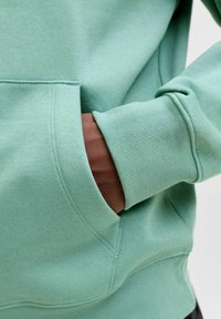 Mint zelena dukserica s teksturiranim finišem, koja ima kenguruhu džep, rebraste manžete i donji rub, te opušteni kroj.