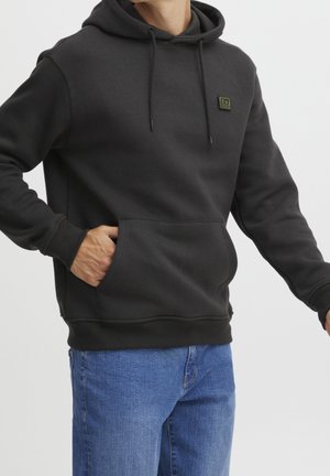 Un homme portant un sweat à capuche noir avec une poche avant et un jean bleu, la main reposant à l'intérieur de la poche, devant un fond clair.