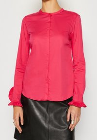 Mujer vistiendo una blusa larga de manga roja vibrante con puños con volantes, combinada con una falda de cuero negra, mostrando la parte superior del cuerpo y las manos.