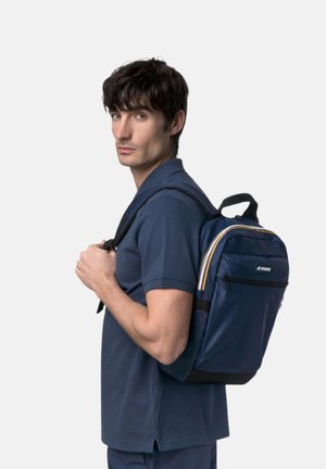 Uomo con camicia blu che indossa uno zaino blu navy con dettagli a righe, che guarda oltre la spalla su uno sfondo semplice.