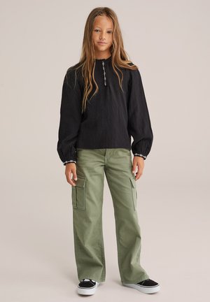 Maglia nera a maniche lunghe con scollo rotondo e cuciture bianche, abbinata a pantaloni cargo verdi con tasche laterali e una vestibilità ampia.