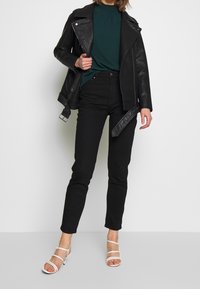 Donna in giacca di pelle nera sopra un top verde a fantasia, jeans neri a vita alta e sandali con tacco bianchi con cinturini, in piedi davanti a uno sfondo bianco.