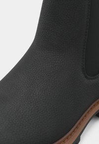 Melns gumijas zābaks ar fakturētu virsmu, elastīgām sānu panelēm un brūnu zoles ārpusi. Raksturots ar apaļu purngalu un šūšanas detaļām.