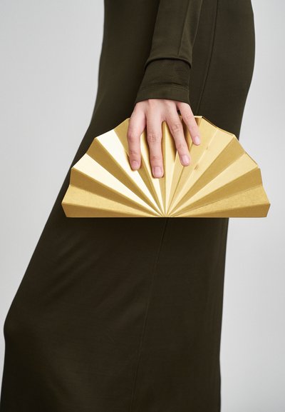 Χρυσή τσάντα χειρός σε σχήμα ανεμιστήρα, με πλισέ σχέδιο και λεία υφή. Κρατιέται με το χέρι μπροστά από φόντο πράσινο φόρεμα.