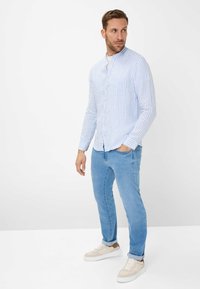Camicia button-up a righe blu chiaro e bianco con maniche lunghe, taschino sul petto e orlo arrotondato, abbinata a jeans in denim chiaro e sneakers.