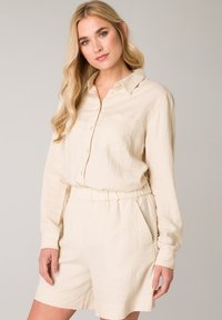 Beige linnen shirtjurk met lange mouwen, knoopsluiting, voorzak en elastische tailleband; lichtgewicht, ontspannen pasvorm en een soepele textuur.