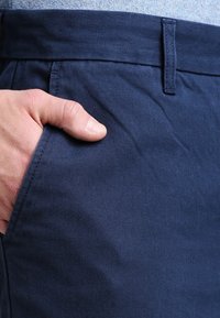 Navy blue trousers with a smooth, textured fabric. Features a waistband and a side pocket with a hand resting in it.