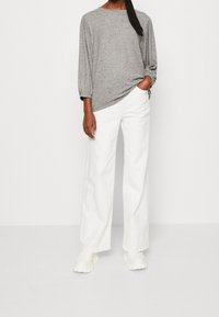 Donna in piedi contro uno sfondo bianco, che indossa un top grigio a maniche lunghe, pantaloni bianchi larghi e scarpe chunky bianche, con una mano in tasca.