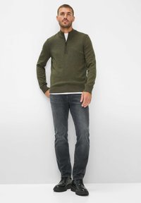 Grüner Reißverschluss-Pullover aus Strickmaterial, kombiniert mit dunkelgrauen Jeans und schwarzen Schuhen. Der Pullover hat gerippte Akzente an den Bündchen und dem Saum.