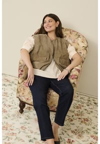 Brun ruskindsvest med to forlommer, båret over en lys bluse og mørke denimjeans, siddende på en stol med blomsterprint og et tæppe.