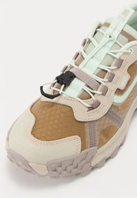 Sneaker beige e marrone chiaro con una tomaia testurizzata, dettagli in rete e suola modellata. Caratterizzato da lacci elastici e un meccanismo di bloccaggio frontale.