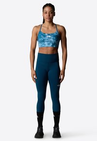 Sujetador deportivo azul con diseño tie-dye, con tirantes delgados, combinado con leggings de talle alto en color azul verdoso. Calcetines negros y zapatos de senderismo completan el look.