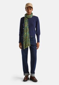 Marineblauer Strickpullover, grüner Blumenschal, blaue Jeans mit hochgekrempelten Säumen und braunen Schuhen mit schwarzen Sohlen. Einfaches, legeres Outfit.