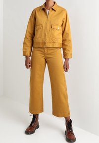 Chaqueta de mezclilla mostaza con dos bolsillos delanteros, diseño corto y pantalones de pierna ancha a juego. Botas marrones con cordones completan el conjunto.