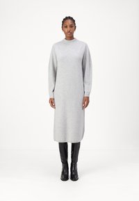 Robe en tricot grise avec un col montant et des manches longues. Présente une silhouette droite, se terminant sous le genou, portée avec des bottes noires hauteur genou.