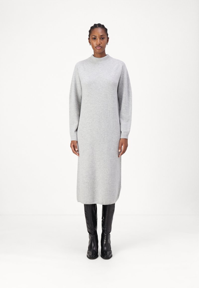 Robe en tricot grise avec un col montant et des manches longues. Présente une silhouette droite, se terminant sous le genou, portée avec des bottes noires hauteur genou.