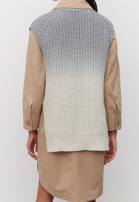 Veste tricotée avec un dégradé du gris au blanc, présentant un design texturé et un ourlet arrière plus long, superposée sur une robe-chemise beige.