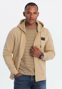 Sudadera marrón clara con cremallera frontal, bolsillos laterales y un parche de logo negro, llevada sobre una camiseta de rayas beige. Combinada con jeans azules.