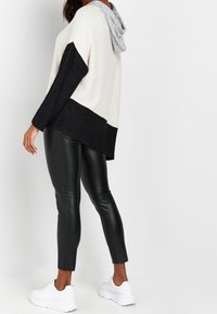 Kvinna iförd en beige och svart färgblockerad hoodie, svarta leggings i faux läder och vita chunky sneakers, står framför en vit bakgrund.