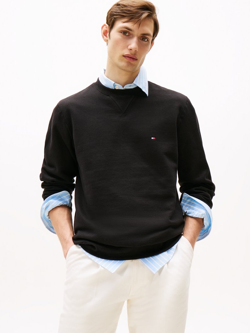 Schwarzer Sweatshirt mit Rundhalsausschnitt, ausgestattet mit einem kleinen Logo. Über einem hellblauen gestreiften Hemd getragen. Weiße Hose, die an der Taille sichtbar ist.