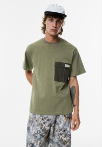 Olivengrøn t-shirt med en mørkegrøn brystlomme, korte ærmer og en afslappet pasform, parret med mønstrede grå shorts og en tofarvet kasket.