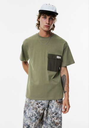 Olivengrøn t-shirt med en mørkegrøn brystlomme, korte ærmer og en afslappet pasform, parret med mønstrede grå shorts og en tofarvet kasket.