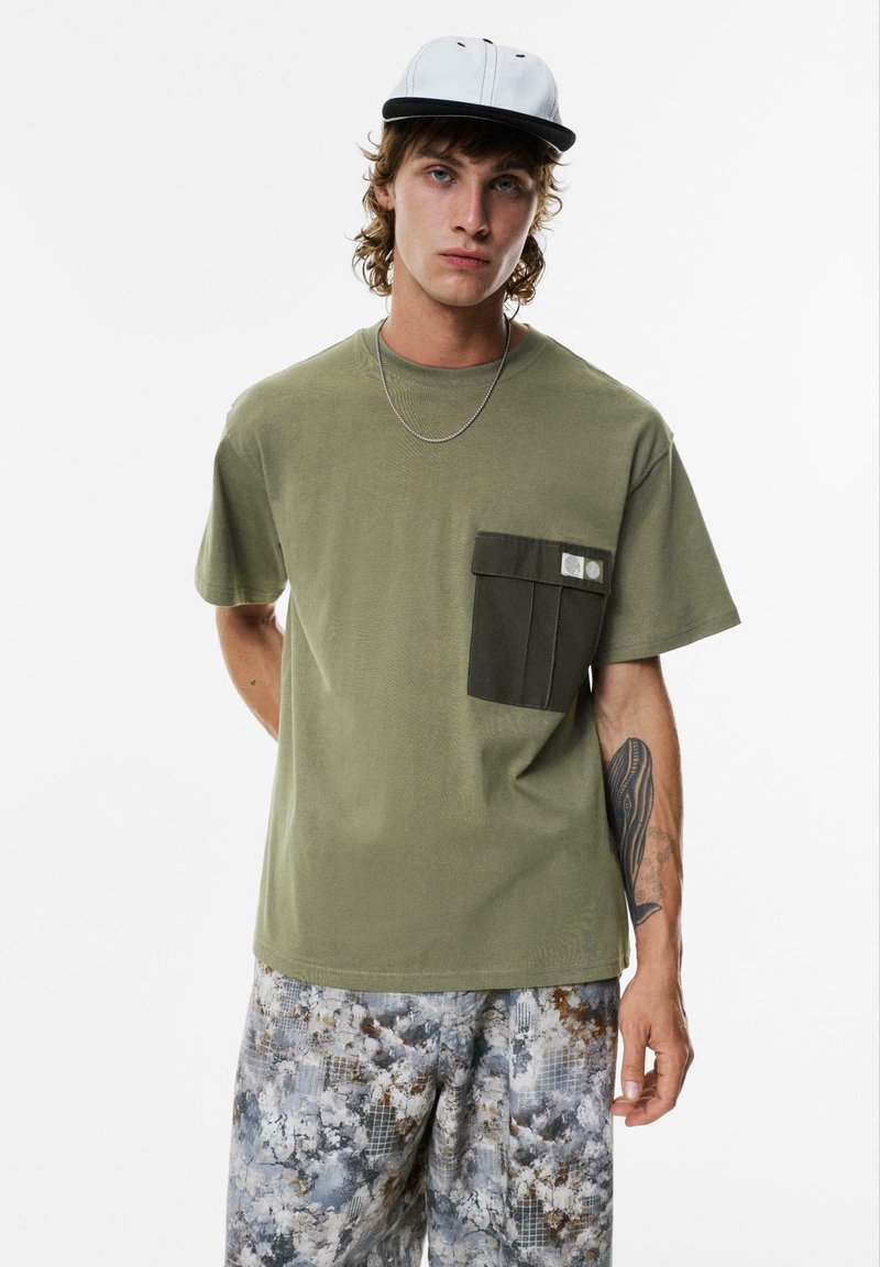 Olivengrøn t-shirt med en mørkegrøn brystlomme, korte ærmer og en afslappet pasform, parret med mønstrede grå shorts og en tofarvet kasket.
