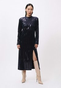 Marineblauwe pailletten jurk met lange mouwen, een hoge halslijn en een zijsplit, gecombineerd met beige glanzende haklaarzen. Smalle textuur, elegante uitstraling.