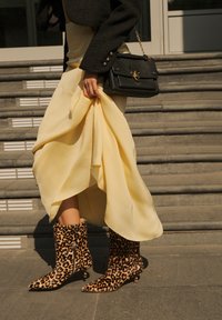 Luipaardprint enkellaarsjes met spitse neuzen en kittenheels, gecombineerd met een vloeiende gele jurk en een gestructureerde zwarte blazer, terwijl een zwarte handtas vastgehouden wordt.