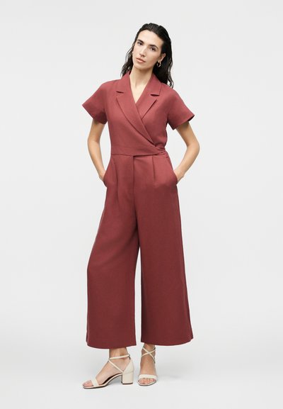 Γυναίκα με σκούρα μαλλιά που φοράει ολόσωμη φόρμα με κοντά μανίκια σε χρώμα σκουριάς και μπεζ σανδάλια με λουράκια και τακούνι μπλοκ, στέκεται με τα χέρια στις τσέπες.