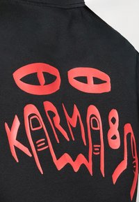 Black t-shirt featuring bold red graphic of eyes and hands with the text "KARMA" in stylized lettering, showcasing a smooth texture.