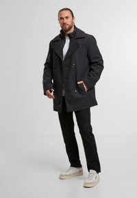 Cappotto peacoat doppiopetto in lana grigia scura con ampi risvolti, tasche laterali e una consistenza morbida, abbinato a pantaloni neri e sneakers bianche.